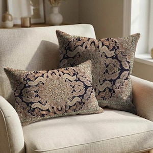May include: Two decorative throw pillows with a dark blue background and an intricate, ornate pattern in shades of beige, brown, and green. The pillows are rectangular and square, placed on a beige armchair.