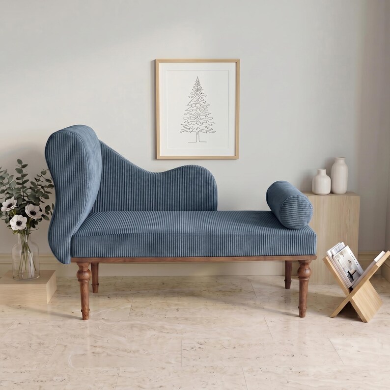 May include: A blue corduroy chaise lounge with a curved back and a cylindrical bolster pillow. The wooden frame has turned legs. A framed line drawing of a tree hangs on the wall behind the chaise lounge.