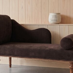 May include: A dark brown corduroy chaise lounge with a curved back and armrest. The wooden frame has turned legs. A small decorative clock and a wooden duck are on a shelf in the background.