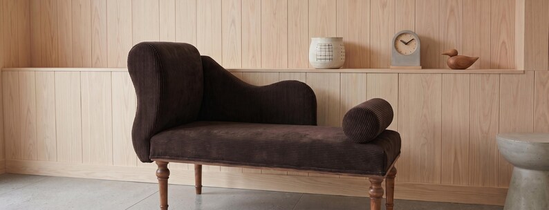 May include: A dark brown corduroy chaise lounge with a curved back and armrest. The wooden frame has turned legs. A small decorative clock and a wooden duck are on a shelf in the background.