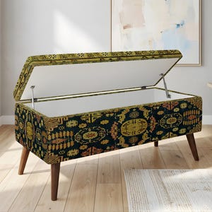 May include: A rectangular storage ottoman with a hinged lid, featuring a dark blue and gold patterned fabric. The ottoman has wooden legs and is open, revealing a white interior. The design is reminiscent of a vintage rug.