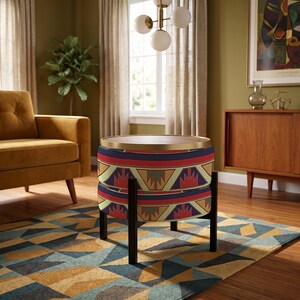 May include: A round ottoman featuring a patterned fabric with geometric shapes in red, blue, yellow, and green. The ottoman has a flat wooden top surface with a gold-toned rim and is supported by four black legs arranged in a cross-base structure.