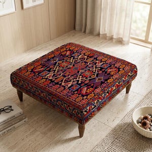 May include: Square ottoman with a patterned rug top in shades of red, blue, and gold. The ottoman has dark wooden legs and is placed on a light-colored tiled floor. A stack of books and a pair of glasses are nearby.