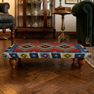 May include: A rectangular footstool with a patterned top in red, blue, yellow, and white geometric designs. It has four wooden legs and sits on a dark wood floor. The footstool is approximately 90 cm long.