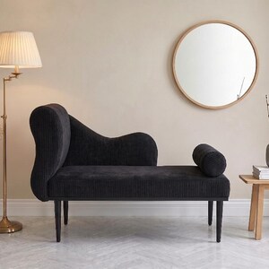 May include: A black corduroy chaise lounge with a curved back and cylindrical bolster. A round wooden mirror hangs on the wall. A brass floor lamp with a pleated shade and a small wooden side table with a vase and books complete the scene.