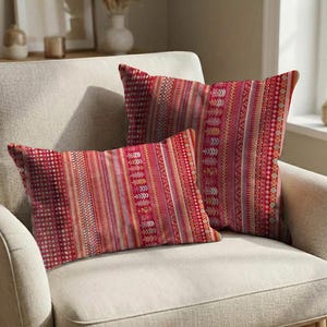 Op de afbeelding: Twee decoratieve sierkussens met een rood-roze patroon. De kussens hebben een mix van verticale strepen en stippenpatronen. De kussens liggen op een beige fauteuil.