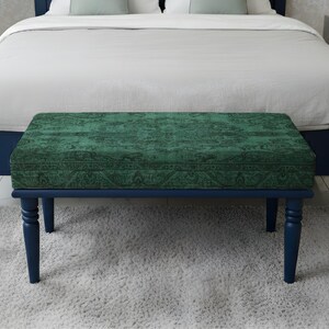 May include: A rectangular bench with a dark teal patterned cushion sits at the foot of a bed. The bench has dark blue legs and frame. The bed has a dark blue frame and white bedding. The bench is on a light grey rug.