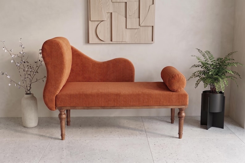 K&ouml;nnte beinhalten: Eine orangefarbene Chaiselongue aus Cord mit geschwungener R&uuml;ckenlehne und zylindrischer Armlehne. Die Holzbeine und der Rahmen tragen die Sitzfl&auml;che. Ein dekoratives Wandkunstwerk aus Holz h&auml;ngt &uuml;ber der Chaiselongue. Eine Vase mit Zweigen und eine Topfpflanze sind ebenfalls im Bild.