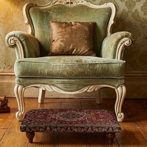 Puede incluir: Un sillón de estilo antiguo con asiento y respaldo de terciopelo verde, adornado con bordados dorados. Cuenta con molduras de madera blanca ornamentadas y un reposapiés a juego con una alfombra roja estampada. Una almohada marrón descansa sobre la silla.