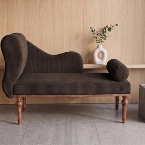 May include: A brown chaise lounge with a high back and a cylindrical bolster pillow. The furniture has a wooden frame and legs. A white vase with greenery and a small side table with a gold tray are also visible.