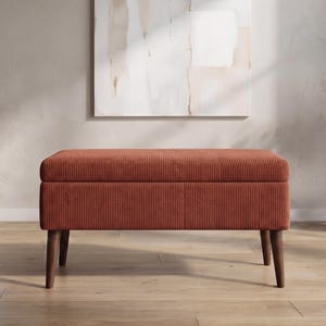May include: A rectangular storage bench upholstered in a rust-colored corduroy fabric with vertical tufted channels. The bench features a hinged lid and is supported by four tapered dark wooden legs that angle outward from the base.