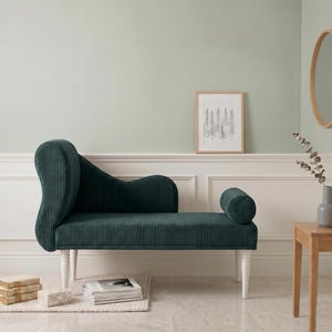 May include: A dark green corduroy chaise lounge with white legs. The chaise has a curved back and a cylindrical armrest. The piece is set against a light green wall, with a framed picture and a small wooden table nearby.