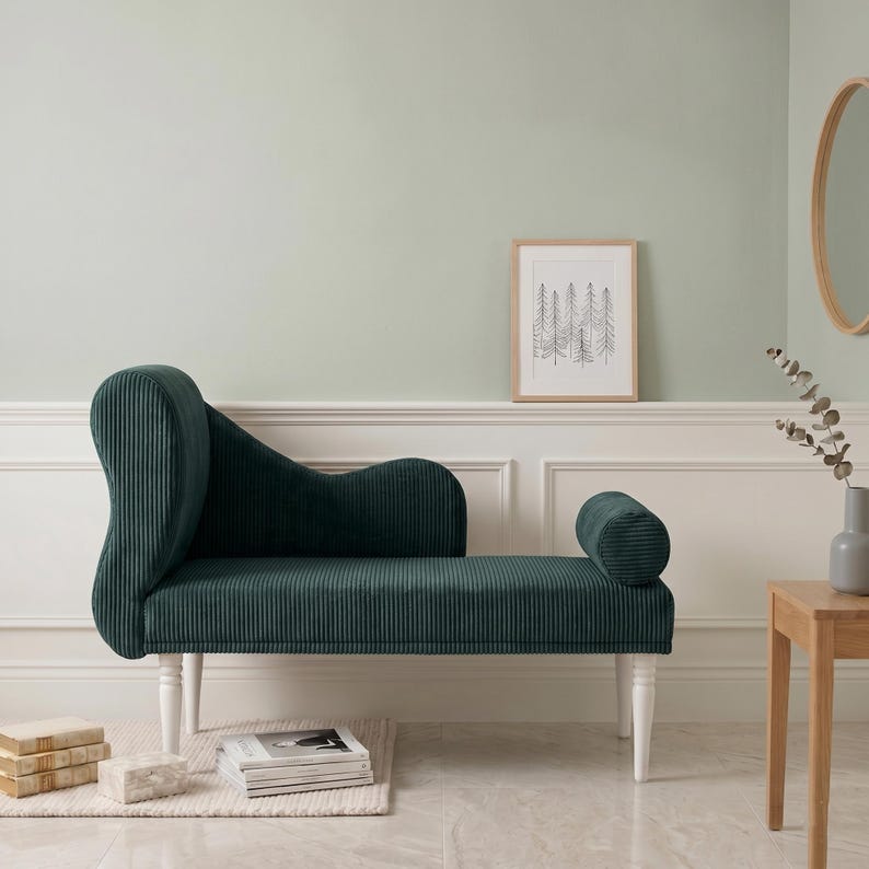 May include: A dark green corduroy chaise lounge with white legs. The chaise has a curved back and a cylindrical armrest. The piece is set against a light green wall, with a framed picture and a small wooden table nearby.