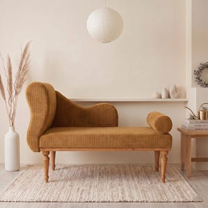 May include: A mustard-colored corduroy chaise lounge with a curved back and armrest. The wooden legs are turned. The chaise is on a woven rug, with a white vase of dried plants to the left.