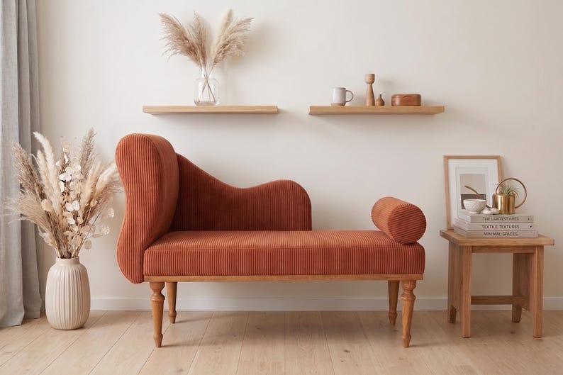 K&ouml;nnte beinhalten: Eine terracottafarbene Chaiselongue mit geschwungener R&uuml;ckenlehne und zylindrischer Armlehne. Der cordartige Stoff ist auf Holzbeinen angebracht. Der Raum ist mit getrockneten Pflanzen, Holzregalen und einem kleinen Beistelltisch dekoriert.