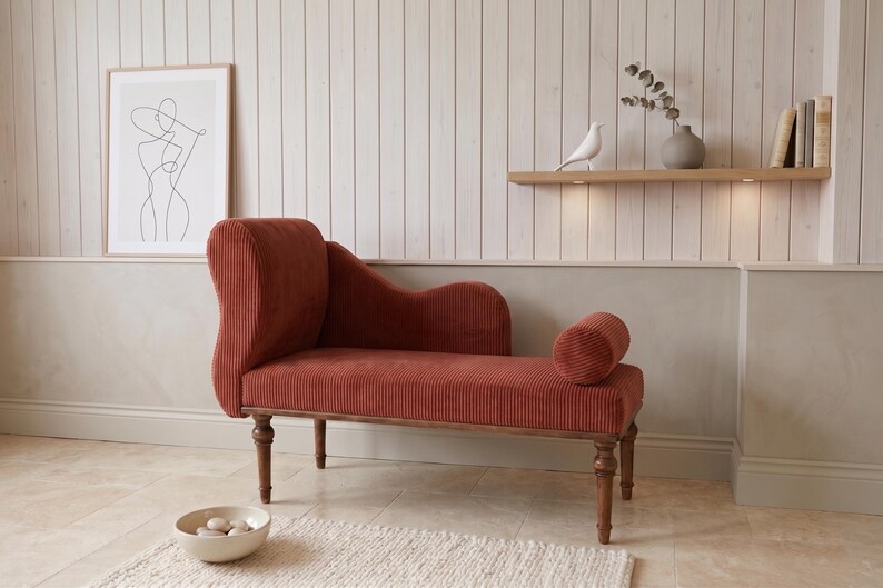May include: A rust-colored chaise lounge with a corduroy texture, wooden legs, and a rolled bolster pillow. The elegant furniture piece is set against a neutral-toned wall with a framed art print and a decorative shelf with a bird figurine.