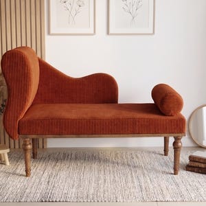 May include: A burnt orange chaise lounge with a high back and a rolled bolster. The corduroy-like fabric is set on wooden legs. A small round mirror and a stack of books are nearby. The chaise is on a neutral rug.