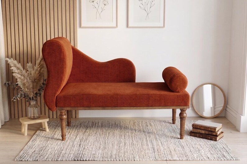 May include: A burnt orange chaise lounge with a high back and a rolled bolster. The corduroy-like fabric is set on wooden legs. A small round mirror and a stack of books are nearby. The chaise is on a neutral rug.