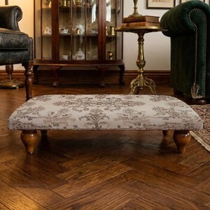Può includere: Un pouf rettangolare imbottito con un motivo floreale beige. Il pouf ha quattro gambe in legno tornito e poggia su un pavimento in legno scuro. Lo sfondo include un divano verde scuro e un mobile in legno.