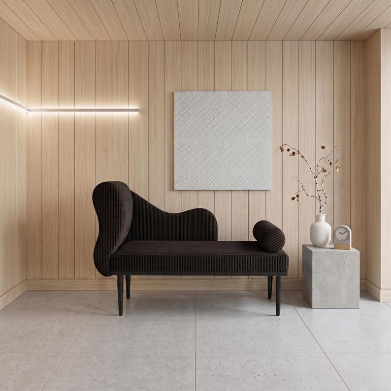 May include: A modern interior scene featuring a dark brown chaise lounge with a curved back and cylindrical bolster. The room has light wood paneling, a white square artwork, and a small side table with a vase and clock.