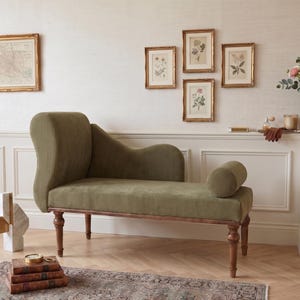 May include: An olive green chaise lounge with a ribbed texture, wooden legs, and a rolled bolster. The room features framed botanical prints, a map, and a vase of flowers. The decor is complemented by a patterned rug and wooden flooring.