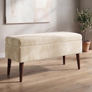 May include: A rectangular storage bench upholstered in a cream-coloured velvet fabric with a smooth, padded top. The bench features four tapered, dark brown wooden legs that provide a stable base for the rectangular seat and storage compartment.