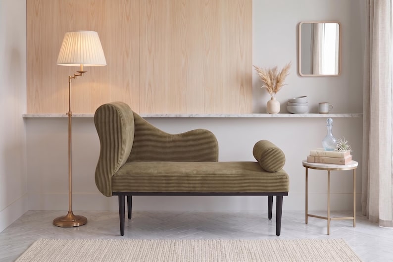 May include: An olive green chaise lounge with a curved back and bolster pillow. A brass floor lamp with a pleated lampshade stands to the left. A small side table and mirror are also in the room.