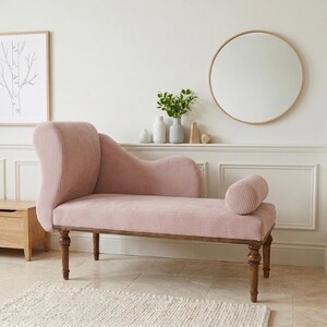 K&ouml;nnte beinhalten: Eine rosa Chaiselongue mit geschwungener R&uuml;ckenlehne und zylindrischem Polster. Die Polsterung hat eine gerippte Textur. Die Holzbeine sind dunkelbraun. Eine Holzaufbewahrungstruhe und eine gerahmte Baumzeichnung befinden sich im Hintergrund.