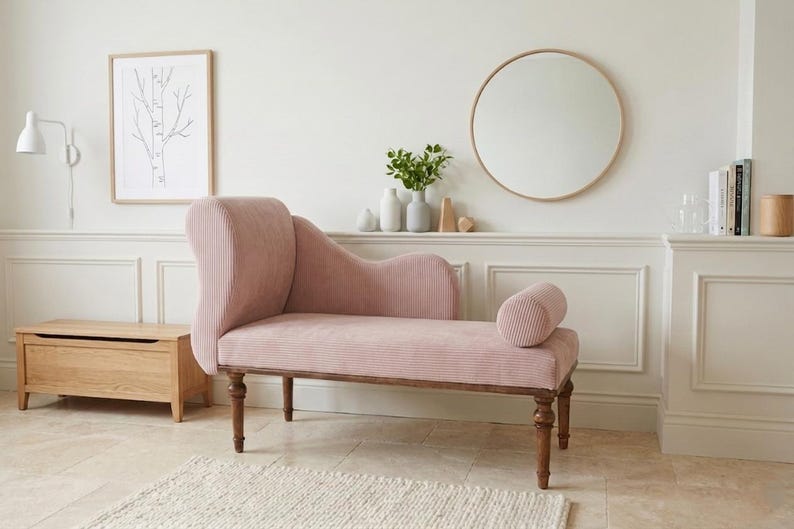 K&ouml;nnte beinhalten: Eine rosa Chaiselongue mit geschwungener R&uuml;ckenlehne und zylindrischem Polster. Die Polsterung hat eine gerippte Textur. Die Holzbeine sind dunkelbraun. Eine Holzaufbewahrungstruhe und eine gerahmte Baumzeichnung befinden sich im Hintergrund.