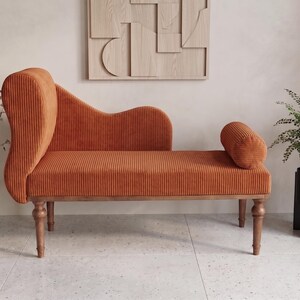 May include: An orange corduroy chaise lounge with wooden legs and a curved back. A cylindrical bolster pillow is on the right side. A vase with branches and a potted fern are on either side.