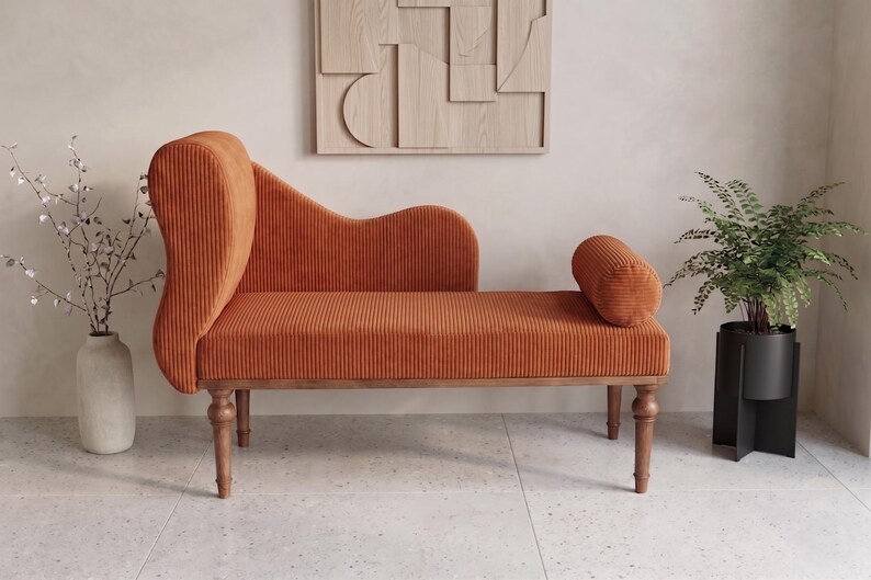 May include: An orange corduroy chaise lounge with wooden legs and a curved back. A cylindrical bolster pillow is on the right side. A vase with branches and a potted fern are on either side.