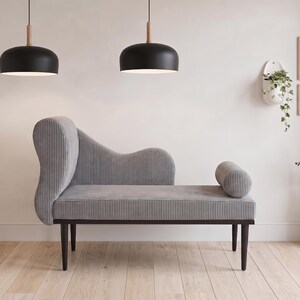 May include: A gray chaise lounge with a curved back and a cylindrical bolster. Two black pendant lights hang above. The room is decorated with framed art, a hanging planter, and a vase with dried flowers. The chaise lounge is approximately 72 inches long.