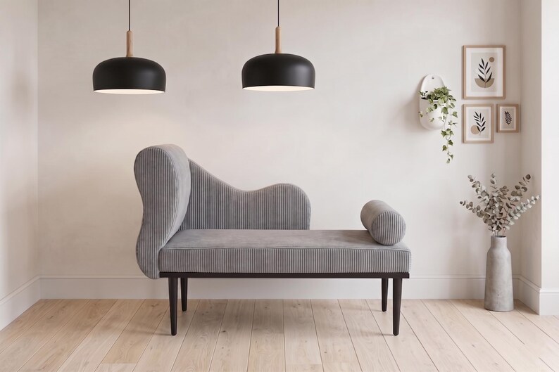 May include: A gray chaise lounge with a curved back and a cylindrical bolster. Two black pendant lights hang above. The room is decorated with framed art, a hanging planter, and a vase with dried flowers. The chaise lounge is approximately 72 inches long.