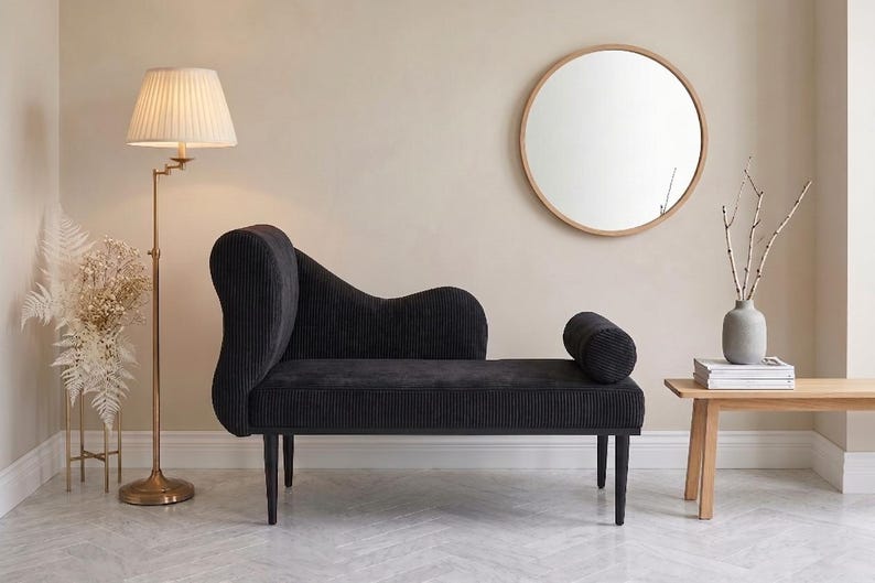 May include: A black corduroy chaise lounge with a curved back and cylindrical bolster. A round wooden mirror hangs on the wall. A brass floor lamp with a pleated shade and a small wooden side table are also present.