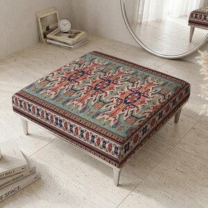 Pode incluir: Pufe quadrado com tampo estampado em tons de verde-azulado, vermelho e azul. O pufe tem uma estrutura de madeira clara e está colocado sobre um chão ladrilhado. Um vaso com flores secas e livros estão ao fundo.