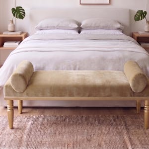 May include: A rectangular bench upholstered in olive green velvet featuring two cylindrical bolster pillows at each end. The bench is supported by four tapered, gold-finished wooden legs, providing a stable base for this piece of furniture.