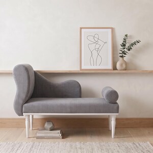 May include: A gray chaise lounge with a ribbed texture and white legs. The furniture piece has a curved back and a cylindrical armrest. A framed line art print, a vase with greenery, and a round mirror are also visible.
