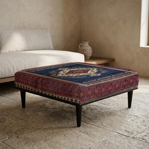 Peut inclure: Un pouf carré avec un dessus à motifs bleu foncé et rouge, rappelant un design de tapis traditionnel. Il a un cadre en bois noir et quatre pieds effilés. La pièce a une palette de couleurs neutres. Le pouf mesure environ 91 cm de côté.