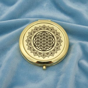 May include: A gold-colored, round compact mirror with a detailed black floral design on the top. The design features a central geometric pattern surrounded by a dotted border and a floral outer edge. The mirror is closed and sits on a blue velvet surface.