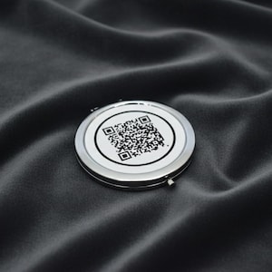 Op de afbeelding: Een ronde, zilverkleurige compacte spiegel met een witte voorkant met een zwarte QR-code. De compact is geplaatst op een donkere, getextureerde stof, wat een contrast creëert tussen het glanzende metaal en het matte oppervlak.