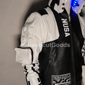 May include: A black and white jacket featuring a high collar with blue LED lighting, the text NUSA printed on the chest, a white sleeve with circular embossed details, a small attached pouch, and a rectangular patch with NUSA text at the hem.