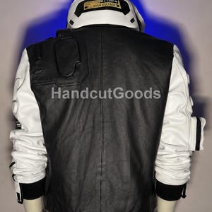May include: A black and white jacket featuring a high collar with a yellow label reading M.2 2280 HIGH VOLTAGE. The back panel is black, while the sleeves are white with black accents and pockets. The text HandcutGoods is visible on the back.