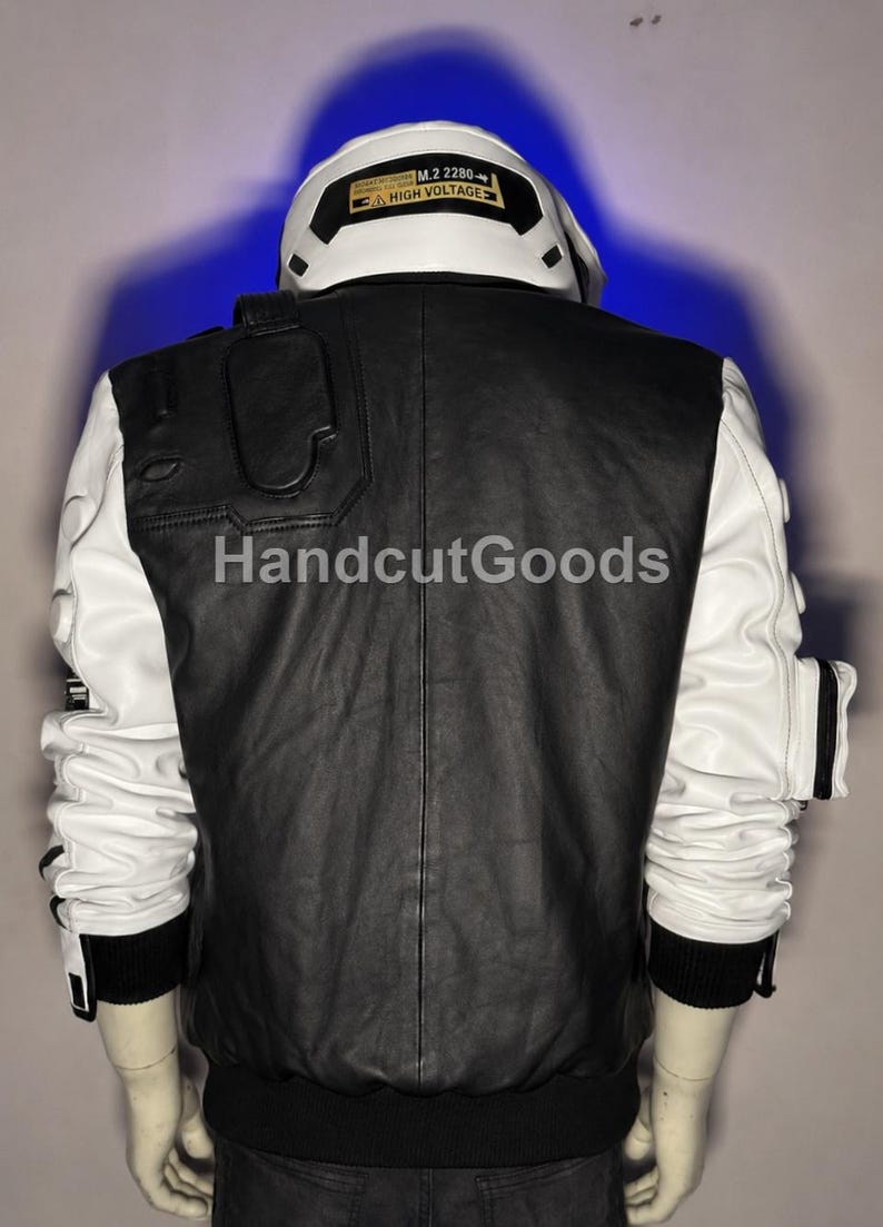 May include: A black and white jacket featuring a high collar with a yellow label reading M.2 2280 HIGH VOLTAGE. The back panel is black, while the sleeves are white with black accents and pockets. The text HandcutGoods is visible on the back.
