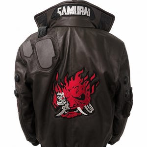 Puede incluir: Una chaqueta tipo bomber de cuero marr&oacute;n oscuro con un gran gr&aacute;fico bordado en rojo y negro de una m&aacute;scara de demonio estilizada en la espalda. El cuello muestra la palabra SAMURAI en letras blancas. Incluye detalles acolchados en hombros y brazos.
