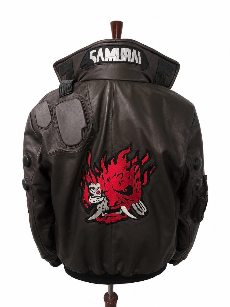 Puede incluir: Una chaqueta tipo bomber de cuero marr&oacute;n oscuro con un gran gr&aacute;fico bordado en rojo y negro de una m&aacute;scara de demonio estilizada en la espalda. El cuello muestra la palabra SAMURAI en letras blancas. Incluye detalles acolchados en hombros y brazos.