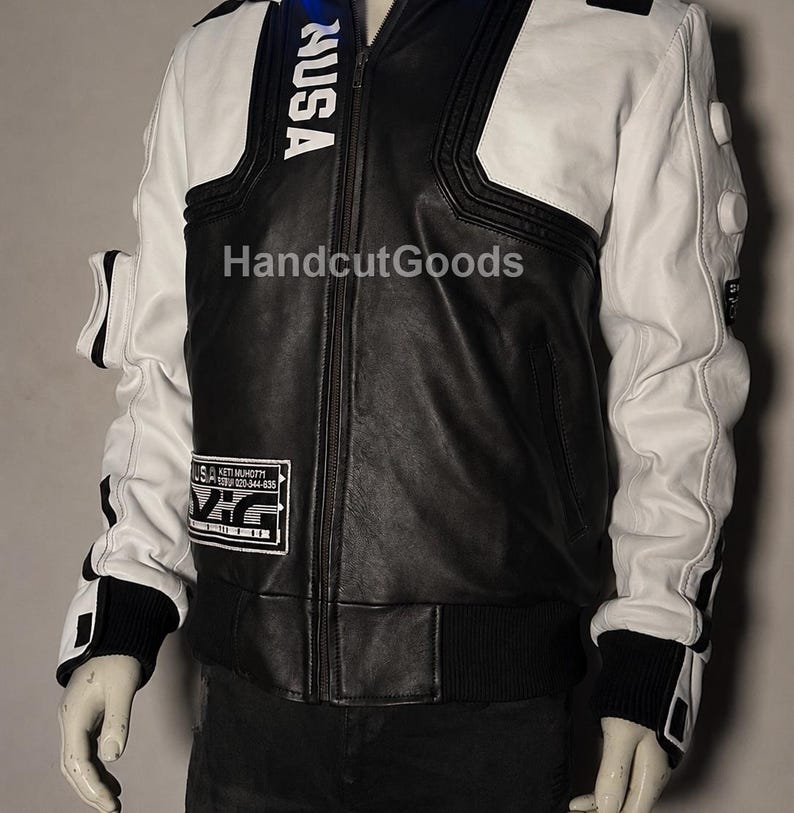 May include: A black and white leather jacket featuring a central zipper, ribbed cuffs, and white sleeves with circular accents. The text NUSA is printed vertically on the chest, and a rectangular patch with technical text is on the lower right side.