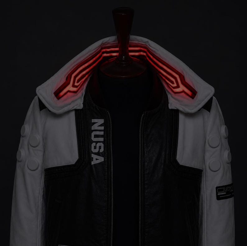May include: A black and white jacket featuring the text NUSA printed vertically on the chest. The garment includes a high collar with glowing red linear accents, three circular buttons on each sleeve, and a dark central panel with a zipper.