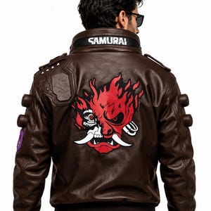 Puede incluir: Una chaqueta de cuero marr&oacute;n oscuro con un gran gr&aacute;fico rojo y negro de una m&aacute;scara de samur&aacute;i rob&oacute;tica estilizada en la espalda. El cuello muestra el texto SAMURAI en letras blancas. La chaqueta incluye charreteras y detalles circulares.