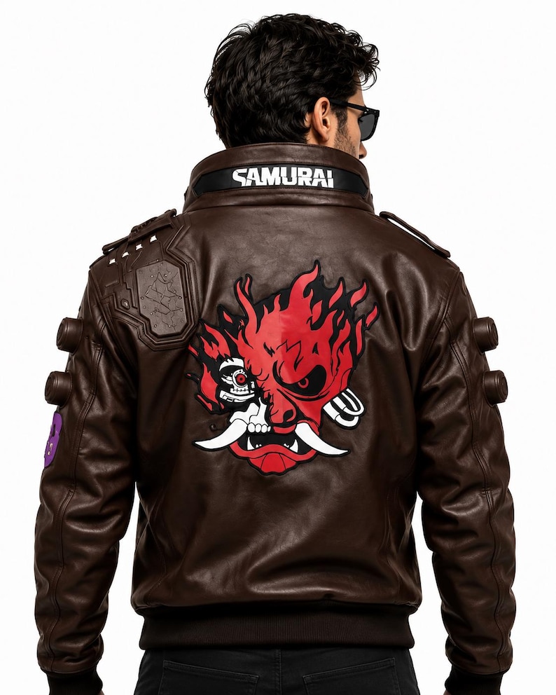 Puede incluir: Una chaqueta de cuero marr&oacute;n oscuro con un gran gr&aacute;fico rojo y negro de una m&aacute;scara de samur&aacute;i rob&oacute;tica estilizada en la espalda. El cuello muestra el texto SAMURAI en letras blancas. La chaqueta incluye charreteras y detalles circulares.