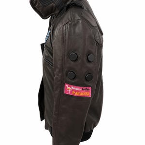 Puede incluir: Una chaqueta de cuero marr&oacute;n oscuro con cuello alto, pu&ntilde;os acanalados negros y cuatro detalles circulares negros en la manga. Un parche rectangular rosa en la parte inferior de la manga muestra un gr&aacute;fico de flamenco y el texto Welcome to Pacifica PARADISE.